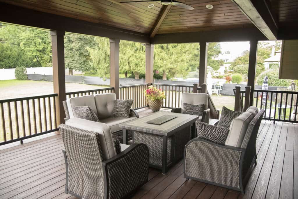 A covered outdoor patio with a wooden ceiling and floor, featuring a seating area with cushioned wicker chairs, a square coffee table, and a flower arrangement. Green trees and a lawn are visible in the background.