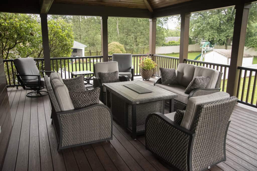 Covered patio with wicker furniture, including a sofa, armchairs, and a fire pit table. Potted plant decorates the table. Green yard, trees, and a children’s playset are visible beyond the dark railing.