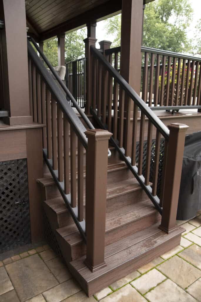 Brown wooden stairs with black railings lead up to a covered porch. The steps and handrails are newly finished, and the area is surrounded by greenery and patio stones.