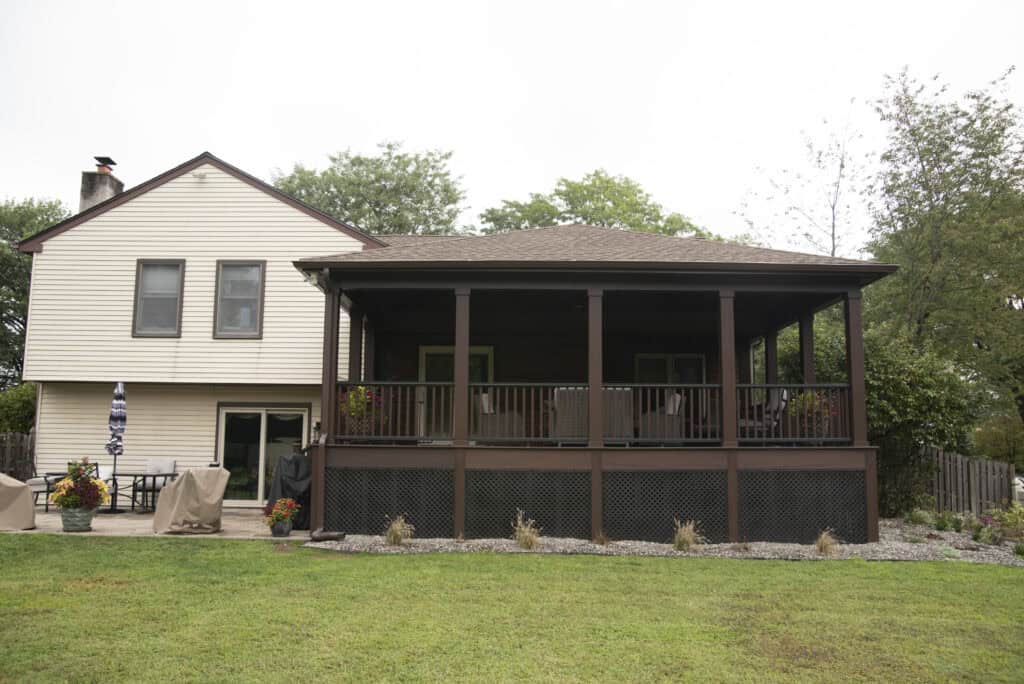 A two-story beige house with a large covered porch featuring dark wood railings and screened panels, overlooking a green lawn and surrounded by trees and plants.
