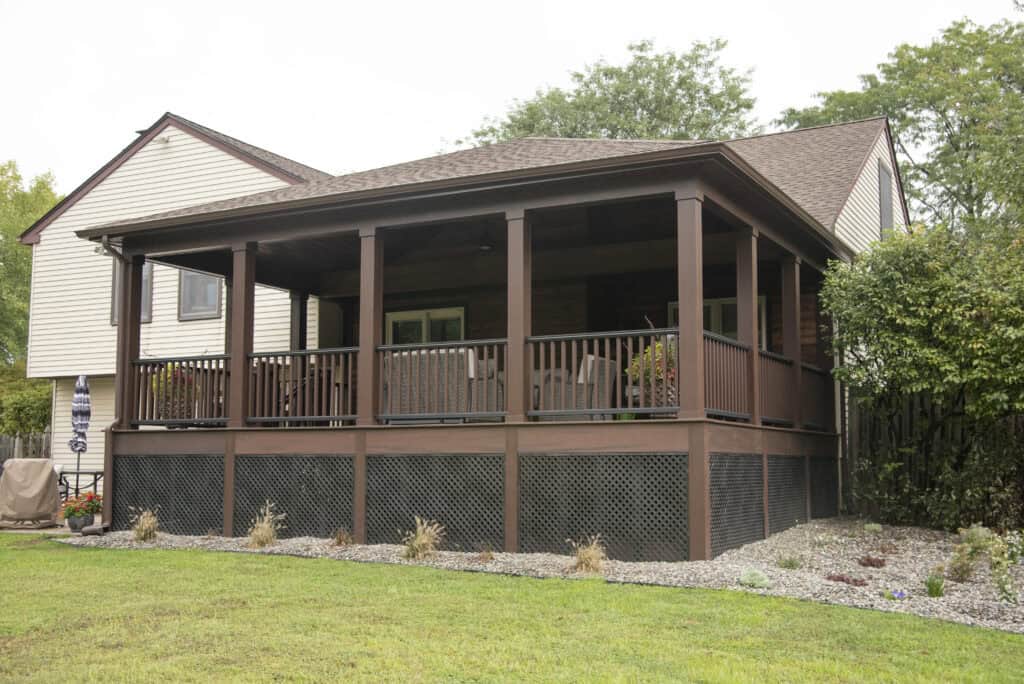 A house with a large covered back porch featuring brown wooden railings and supports, lattice skirting, and a landscaped yard with gravel, small plants, and grass.