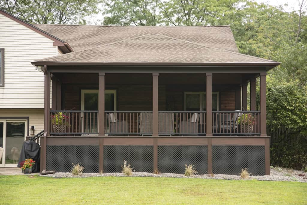 A covered back porch with dark wood railings and columns, featuring outdoor seating and plants, attached to a beige house with a grassy yard in the foreground and trees in the background.