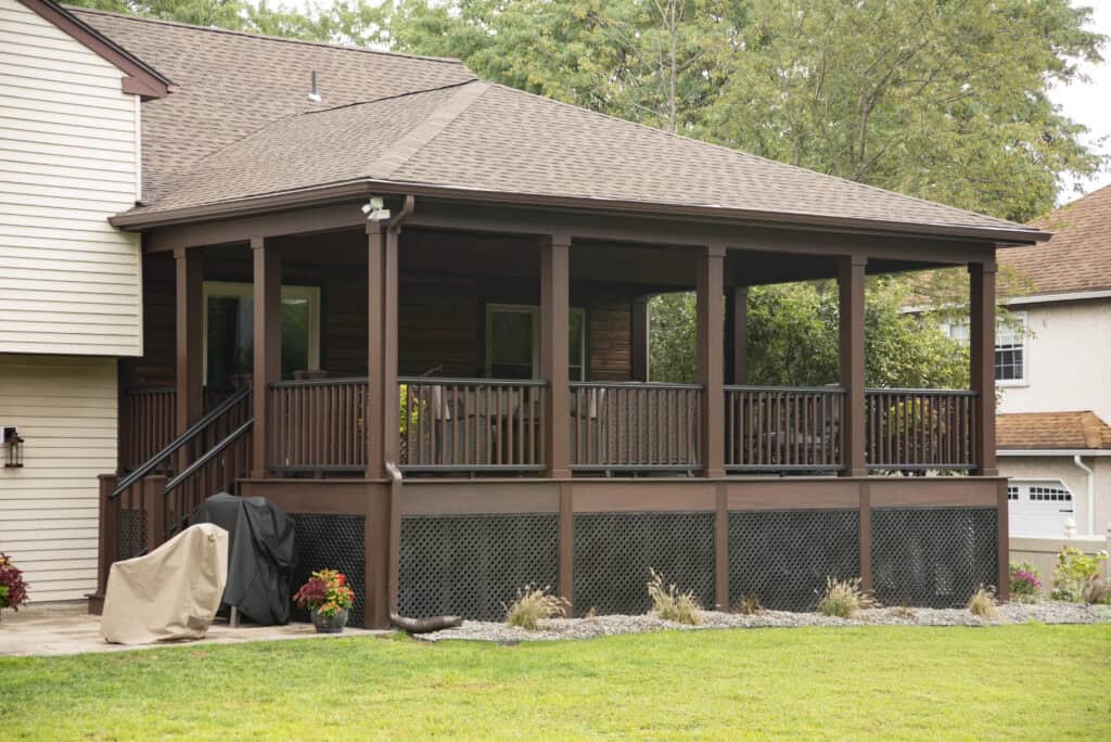 A covered wooden porch with dark railings and lattice skirting is attached to a house. There is patio furniture inside the porch, a grill covered outside, and potted plants lining the edge. Lush green lawn in the foreground.