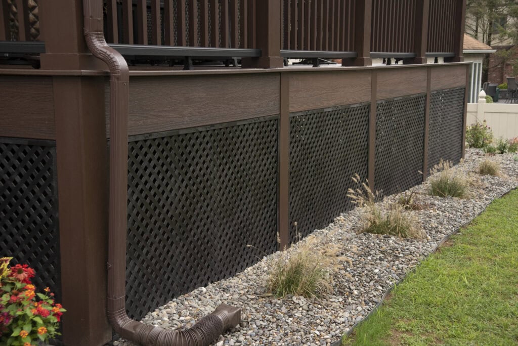 Brown rain gutter and downspout on the side of a raised deck with dark lattice skirting. Decorative rocks and small plants line the base, and a green lawn is visible to the right.