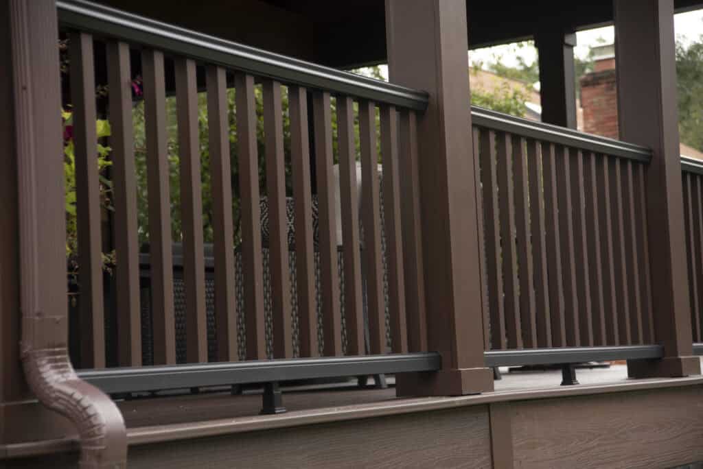 Close-up view of a brown wooden porch railing with vertical slats and black horizontal support bars, attached to a deck with plants visible in the background.