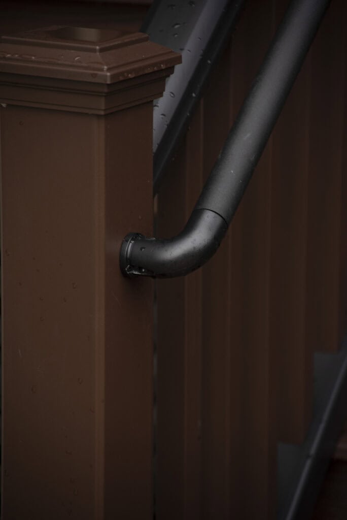 Close-up of a black handrail attached to a brown wooden post on a staircase, with water droplets visible on the surfaces, suggesting recent rain.