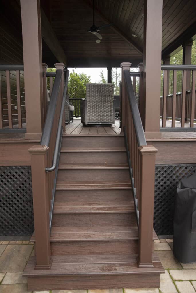 A set of brown wooden stairs with black railings leads up to a covered porch area with outdoor furniture and a ceiling fan. Lattice panels cover the space beneath the porch.