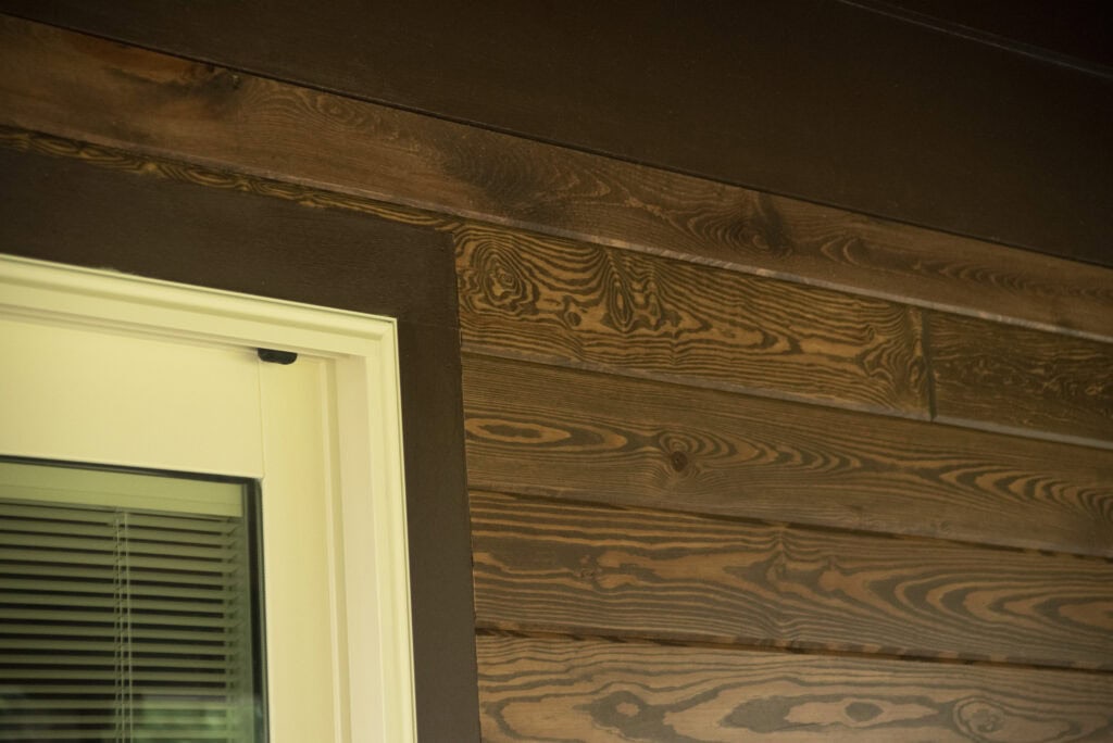 Close-up of a wooden house exterior with dark-stained horizontal wood panels featuring a natural grain pattern. A partially visible window with closed blinds and a white frame is on the left side.