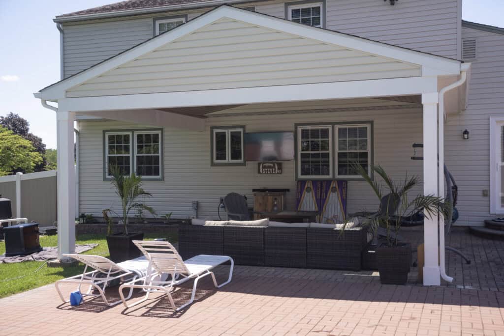 A covered backyard patio with outdoor furniture, potted plants, and two white lounge chairs on a brick surface. The patio is attached to a light-colored house with several windows.