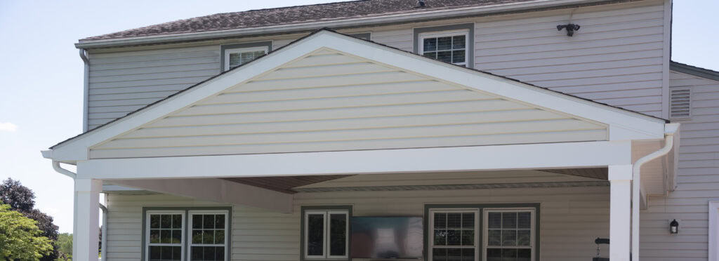 A beige two-story house with white trim featuring a covered patio area. The patio has a white peaked roof supported by columns, and large windows and doors open onto the patio.