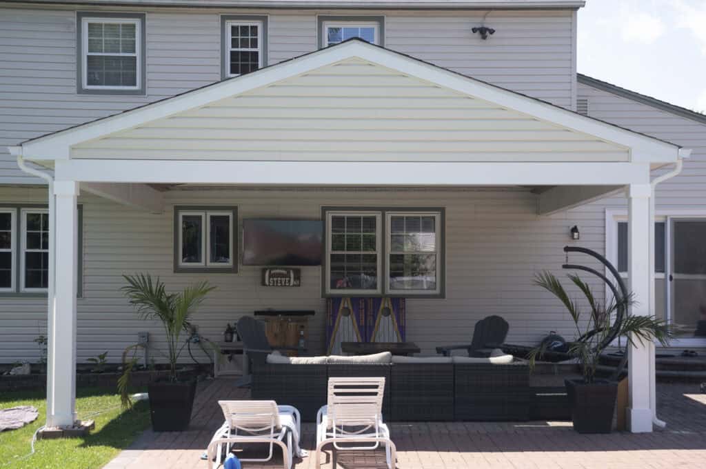 A backyard patio with lounge chairs, an outdoor sofa, armchairs, a TV, and potted plants under a white pergola attached to a light-colored house. Sunlight and shadows suggest a clear day.