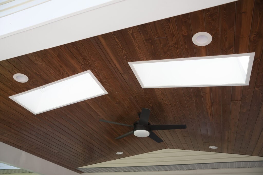 A wooden ceiling with two large rectangular skylights, a modern black ceiling fan with a light in the center, and several round recessed lights.
