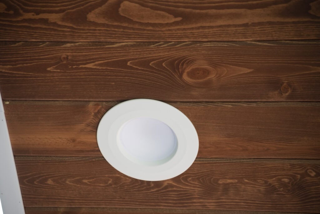 A round recessed ceiling light with a white trim is installed in a wooden ceiling with visible grain and knots.
