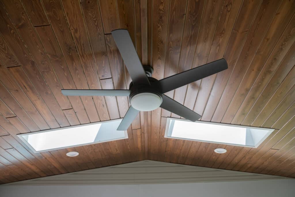 A modern black ceiling fan with five blades is mounted on a wooden ceiling, flanked by two rectangular skylights and recessed lighting.