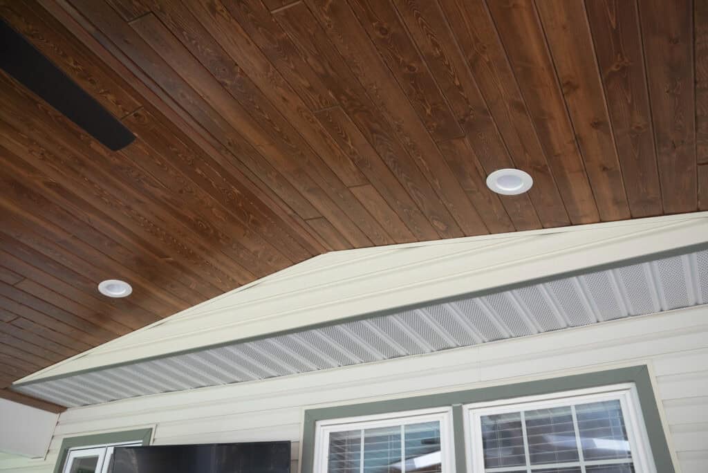 A view of a porch ceiling with rich brown wooden planks, recessed lighting, white siding, and green-trimmed windows below. The design features an angled transition between the wood and white ceiling areas.