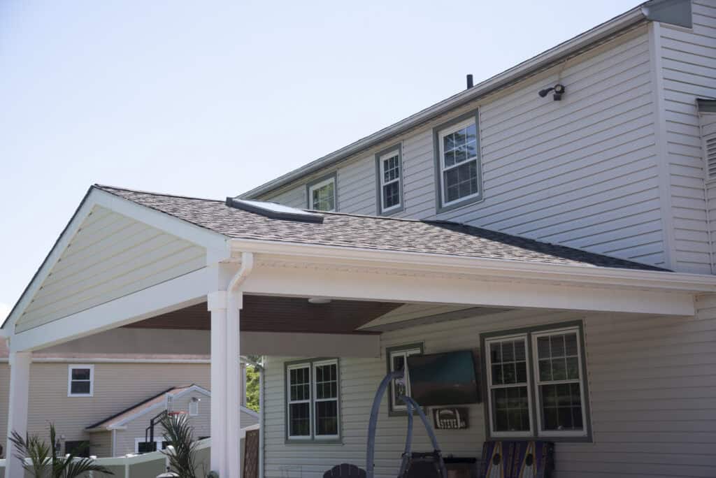 A beige house with white trim features a covered patio area, multiple windows, and outdoor seating. Bright sunlight casts shadows on the building and surrounding area.