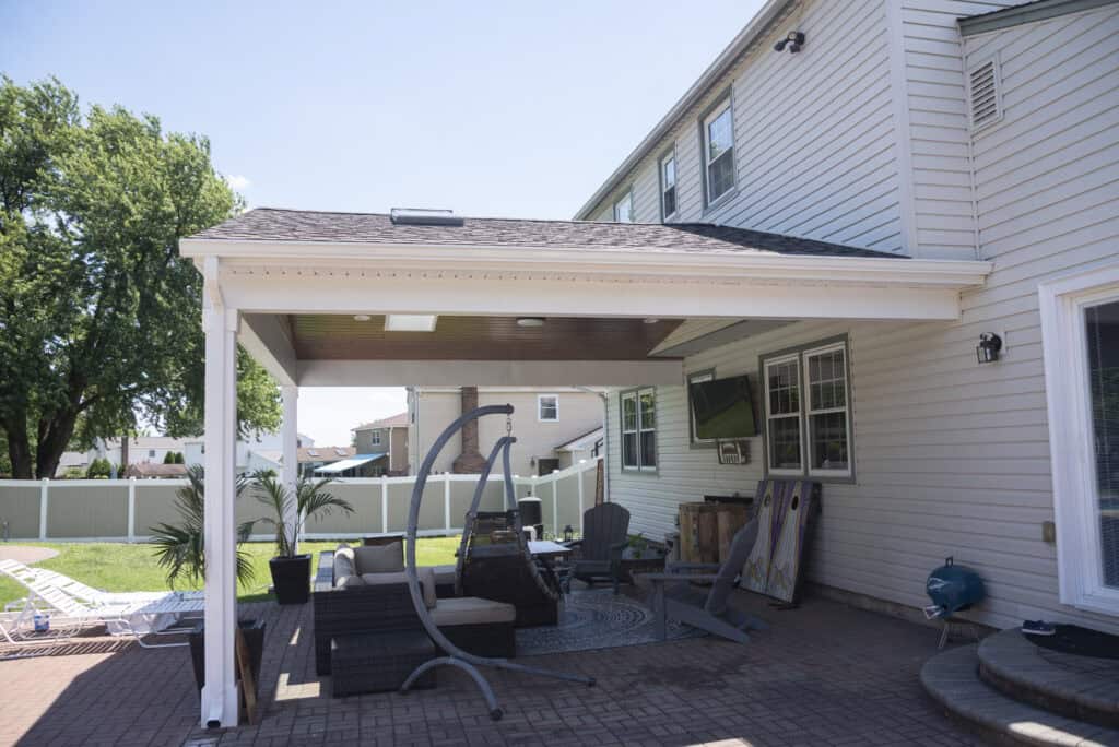 A covered patio attached to a beige house features outdoor furniture, a swing chair, a fire pit, and a TV. The yard has green grass, a fence, and trees visible in the background.