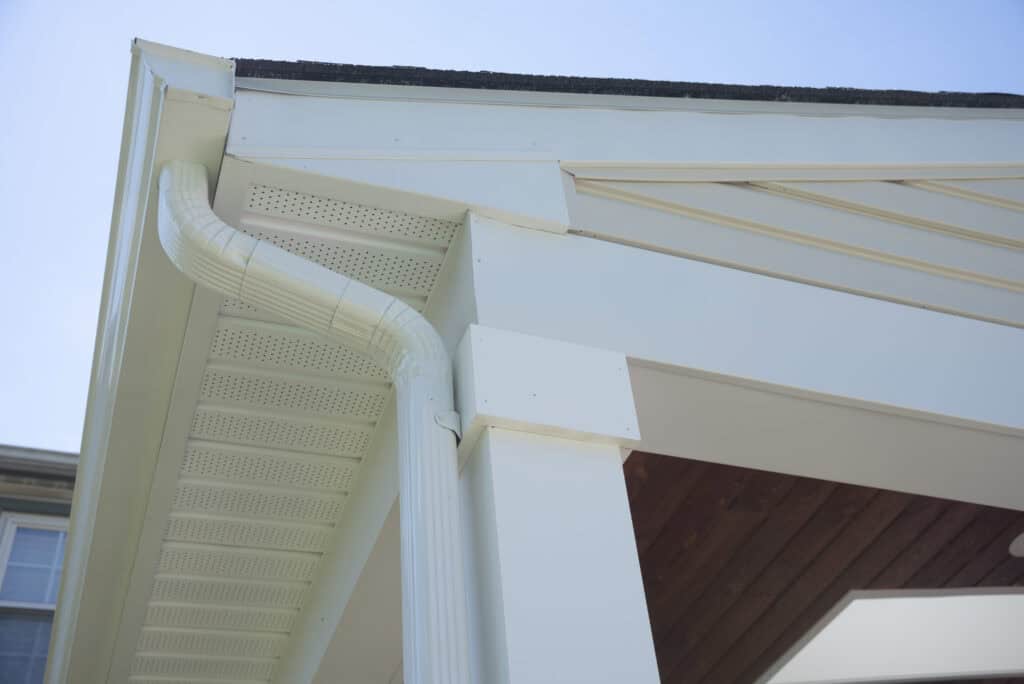 Close-up view of a white house gutter and downspout attached to the roof corner, with a blue sky in the background and part of a wooden porch ceiling visible.