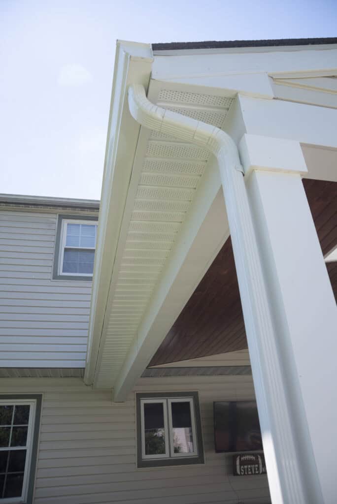 A close-up view of a light-colored house exterior showing a gutter system along the roof edge, with a downspout curving down the side of a covered porch area. Part of the house, roof, and windows are visible.