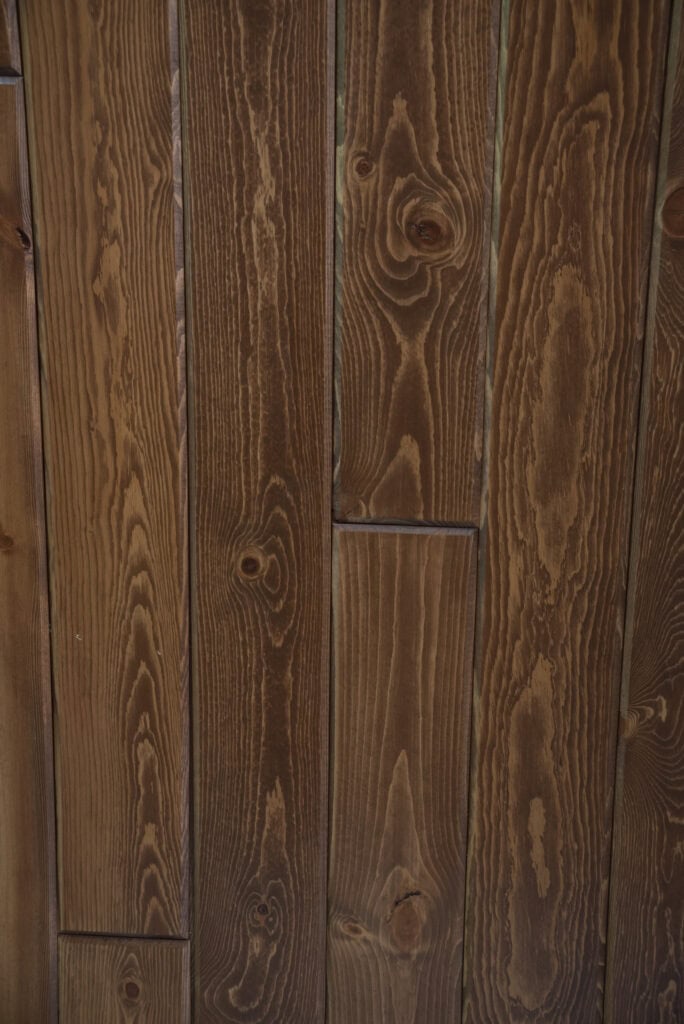 Close-up view of a wooden wall made of vertically aligned planks with visible grain patterns and knots, stained in a warm brown color.