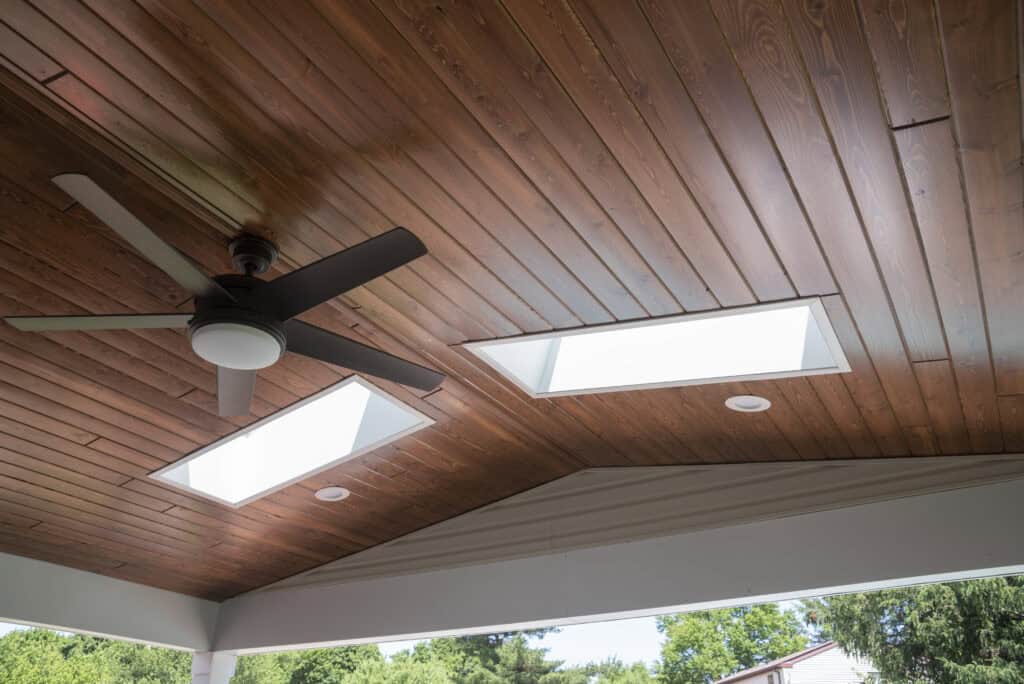 A wooden ceiling features two rectangular skylights and a dark ceiling fan with five blades. Trees and rooftops are visible outside below the ceiling.