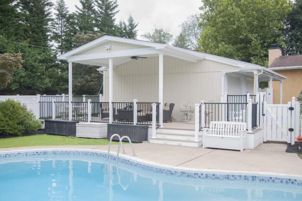 A backyard view of a covered patio with outdoor seating, white railing, and steps leading down to a swimming pool. Trees and shrubs surround the area, creating a peaceful and inviting atmosphere.