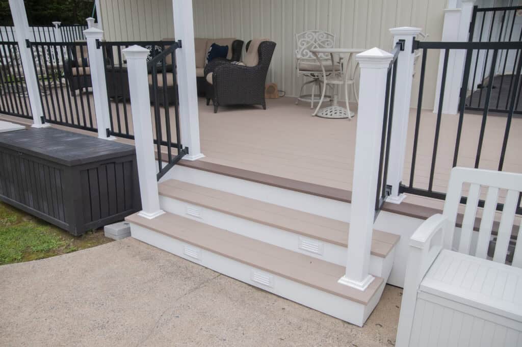 Three tan steps with white trim lead up to a covered deck with black railings and outdoor wicker furniture. A storage bench is on the left, and a white chair is on the right. The area appears clean and well-maintained.