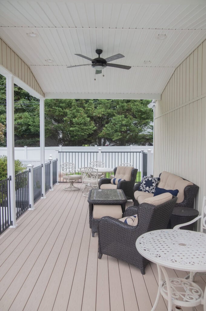 A covered patio with wicker lounge chairs, a cushioned loveseat, metal tables and chairs, ceiling fan, and a view of trees and a white fence in the background.