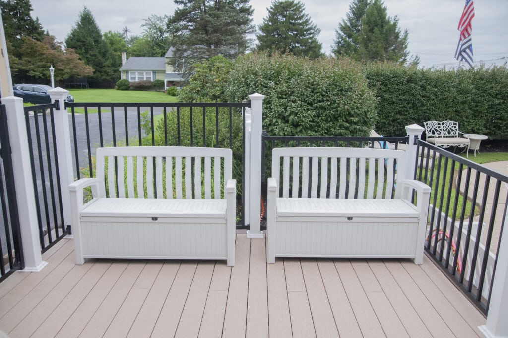 Two white storage benches sit side by side on a light brown deck with black and white railings, overlooking a green yard with trees, a house, and an American flag in the background.