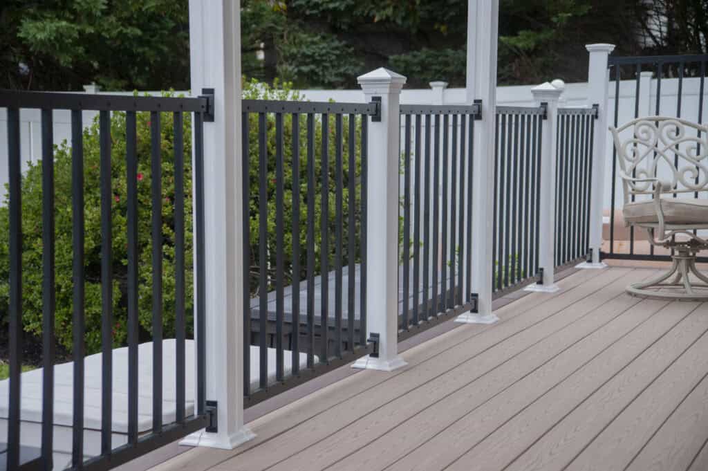 A close-up view of a deck with light brown wood-like flooring, featuring black metal and white posts railing. Patio chairs are partially visible, and green bushes can be seen in the background.