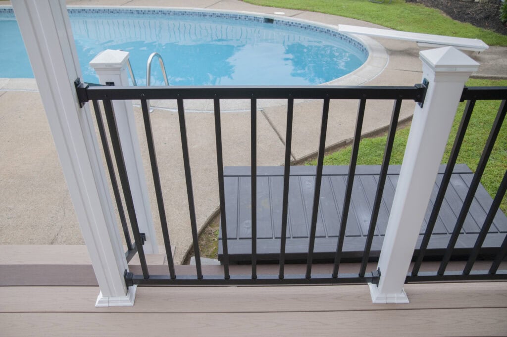 View from a deck with light brown flooring and a black-and-white railing, overlooking a swimming pool, concrete poolside area, green grass, and pool steps.