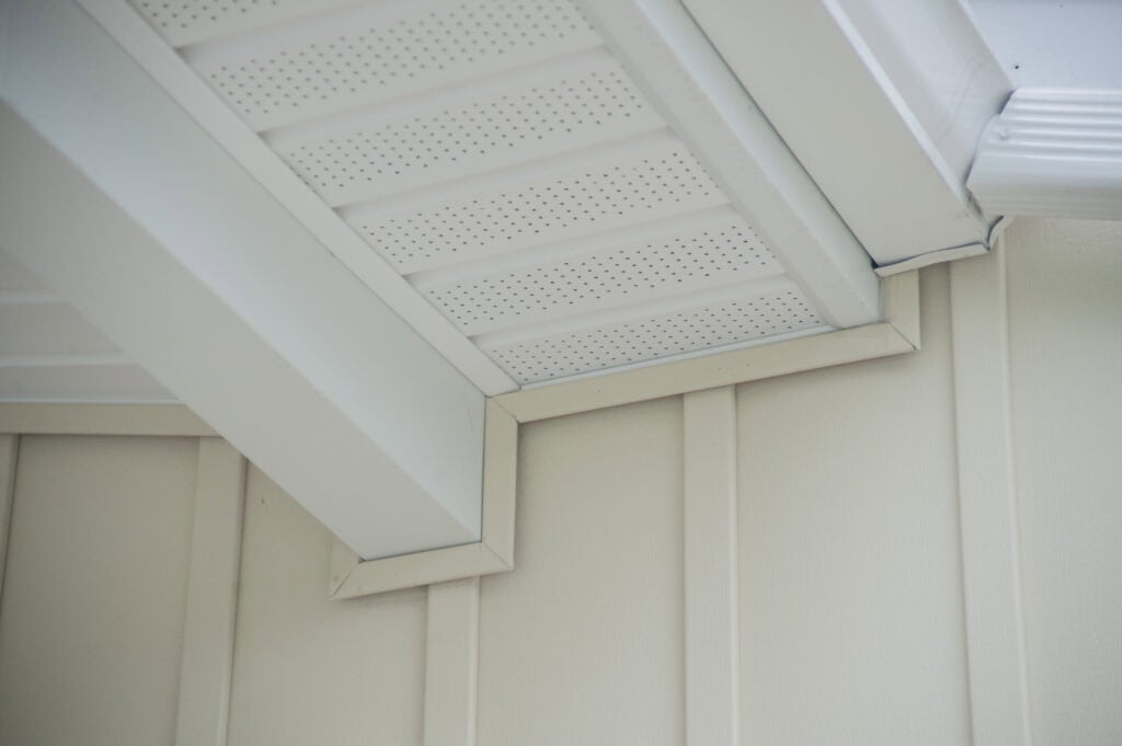 Close-up view of the underside of a house roof showing white soffit panels with small ventilation holes, fascia boards, and beige vertical siding on the exterior wall.