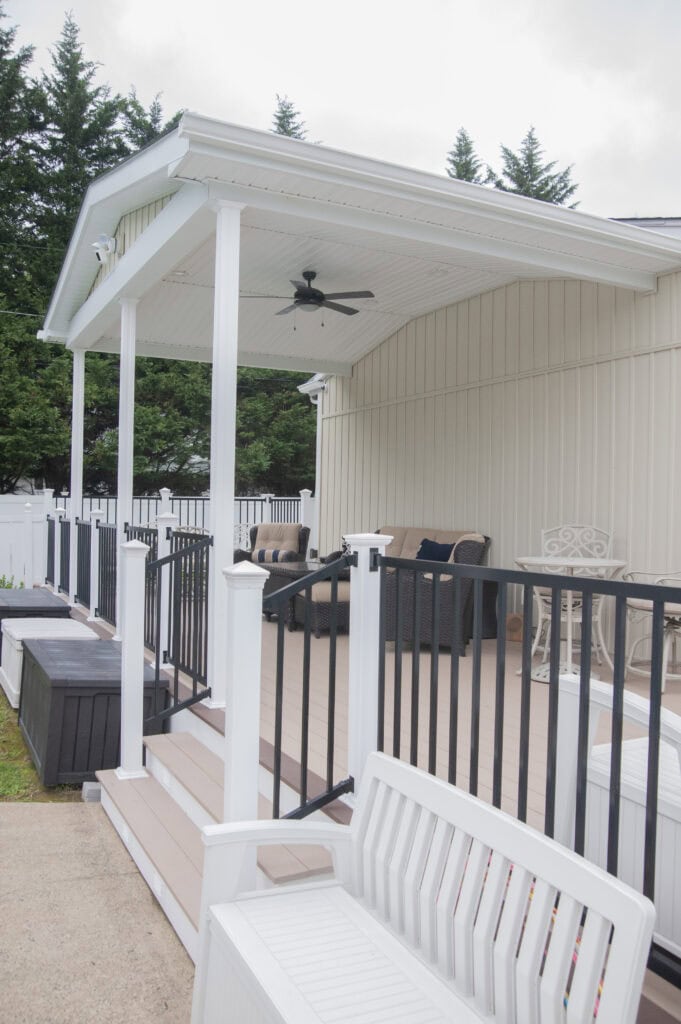 A covered porch with steps, white railings, outdoor seating, and a ceiling fan. The porch is attached to a light-colored building and surrounded by trees and a white fence.