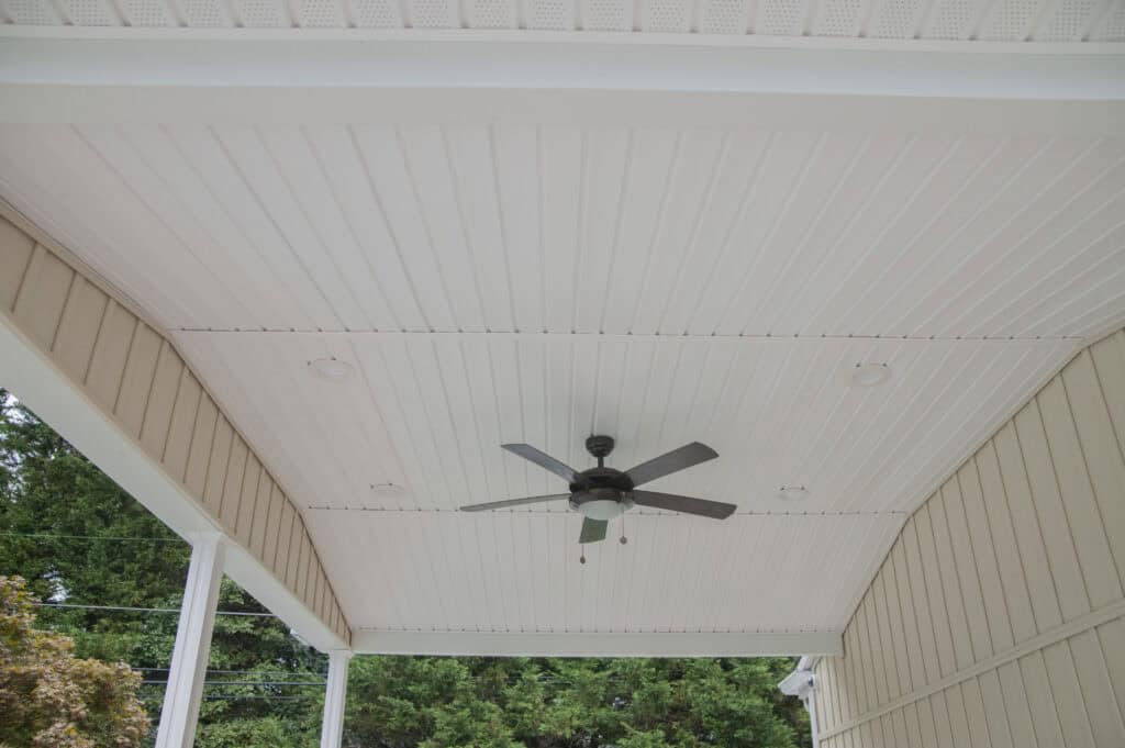 A white beadboard porch ceiling with recessed lights and a black ceiling fan, bordered by beige siding and surrounded by green trees.