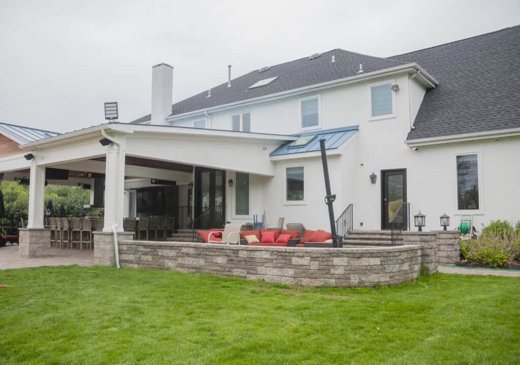 A modern two-story house with white exterior walls, a covered patio area, outdoor seating with red cushions, and a well-maintained green lawn in the foreground.