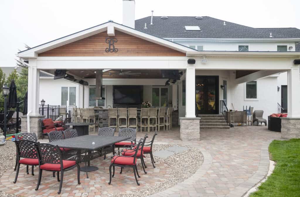 A covered outdoor patio features a long bar with stools, a TV, and a ceiling fan. In front, there is a black metal dining table with red-cushioned chairs on a stone patio, next to a grassy lawn.
