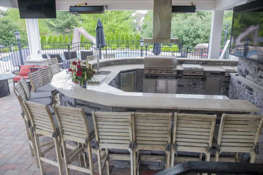 Outdoor kitchen with a curved stone bar, high wooden chairs, stainless steel grill, sink, and countertop. A vase with flowers is on the bar, and patio furniture and greenery are visible in the background.