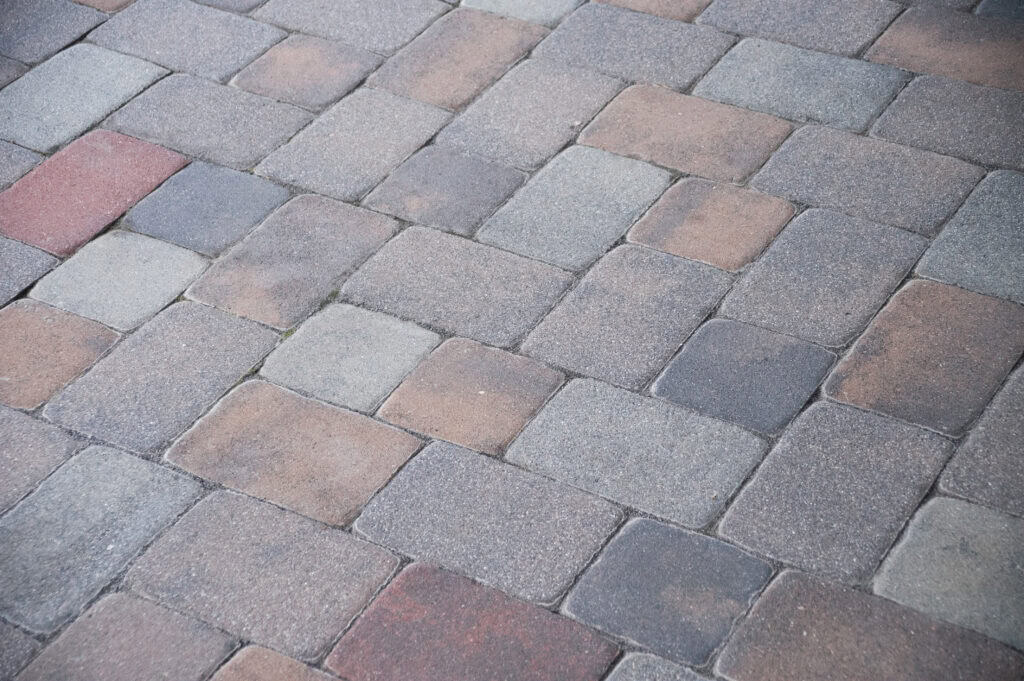 Close-up view of a paved surface made of rectangular and square stone tiles in shades of gray, brown, and red, arranged in a repeating pattern.