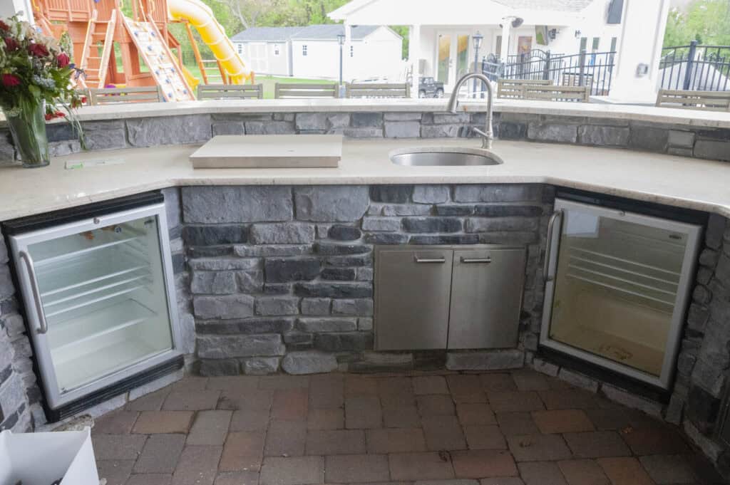 An outdoor kitchen with a stone countertop, featuring a sink, two mini fridges with glass doors, and stainless steel cabinet doors beneath the counter. A playground with a yellow slide is visible in the background.
