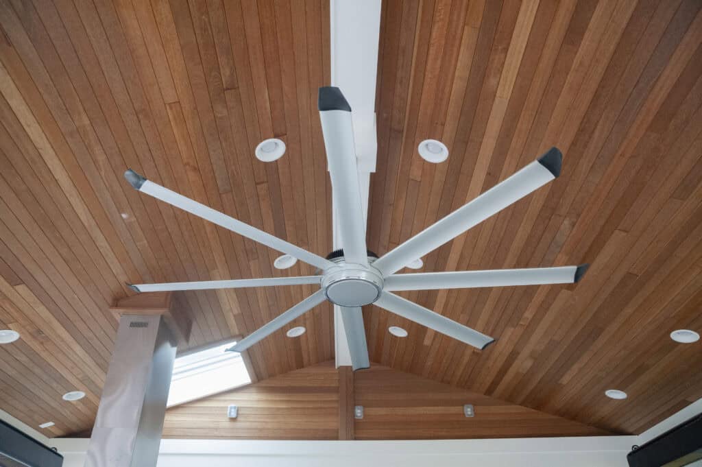 A large silver ceiling fan with eight blades is mounted on a high, wooden-paneled ceiling with recessed lights and a skylight.