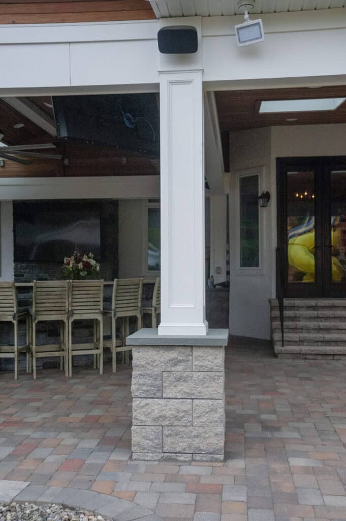 A white column with a stone base stands on a patio with multicolored pavers. Behind it is an outdoor dining area with chairs, a table with flowers, and steps leading to a glass door.