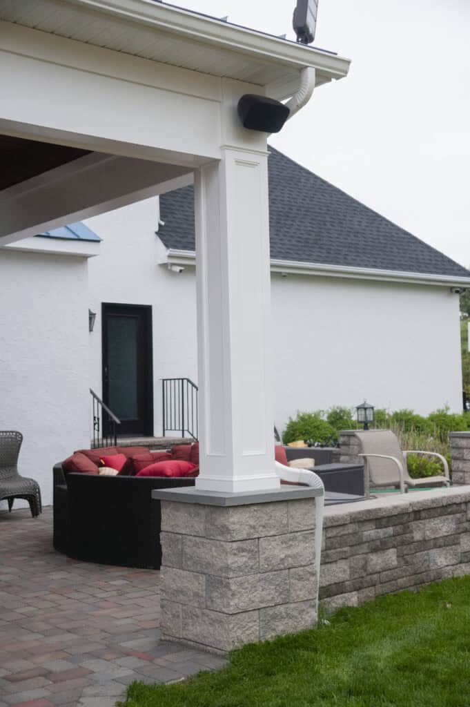 A covered patio with a white column and stone base features outdoor seating with red cushions. The area is paved with bricks and is adjacent to a white house with black-framed doors and windows. Green landscaping surrounds the patio.