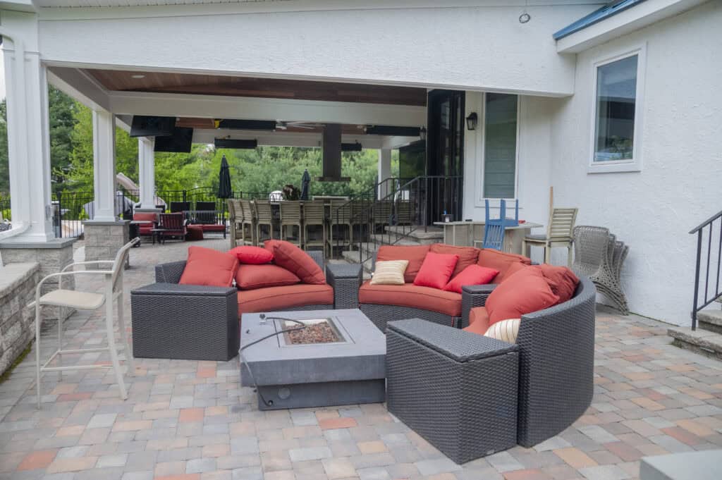 A patio with wicker furniture and orange cushions arranged around a fire pit. In the background, there is an outdoor bar, bar stools, and a covered seating area. The patio surface is paved with multi-colored bricks.