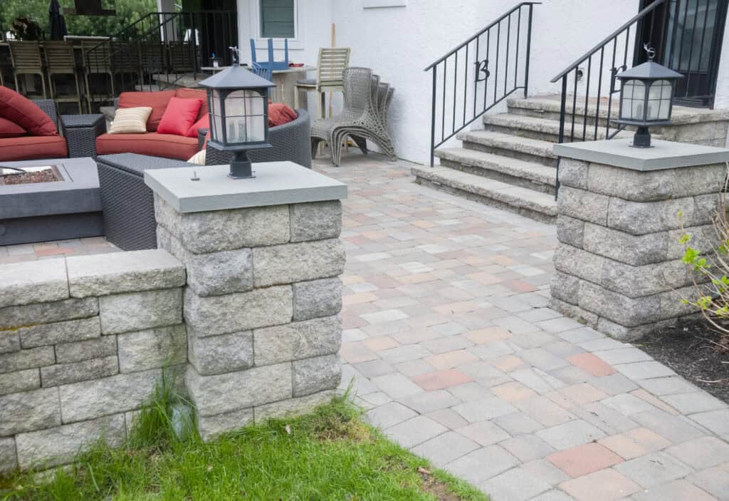 Stone pillars with lanterns frame the entrance to a patio with pavers, outdoor furniture, and steps leading up to a white house. Red cushions and wicker chairs are visible on the patio.