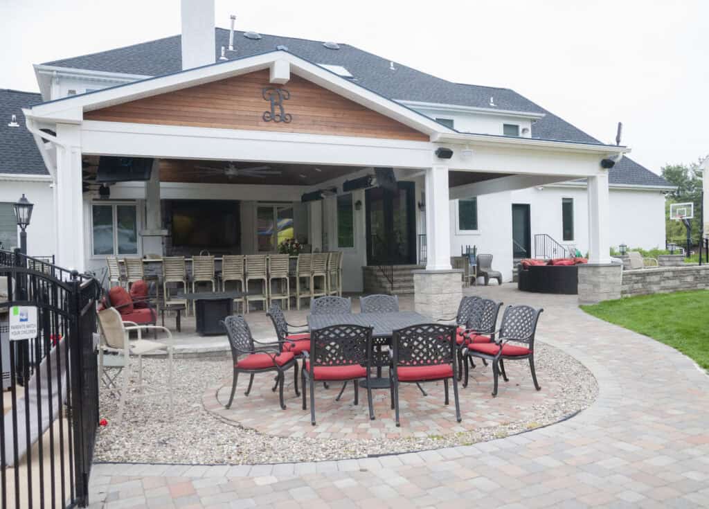 A spacious backyard patio with a covered outdoor bar and kitchen area, barstools, a dining table with red-cushioned chairs, a fire pit, and a basketball hoop in the background.