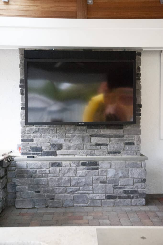 A flat-screen TV is mounted on a stone wall above a narrow stone shelf, with part of a countertop visible at the bottom. The TV screen shows a blurry reflection of a person in yellow.