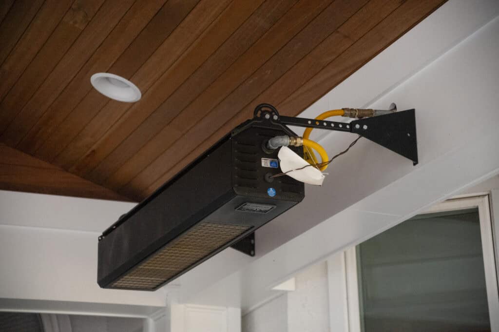 A black rectangular outdoor heater is mounted to a ceiling with a wooden finish. An orange gas hose is connected to the heater, and a white cloth is tied around a pipe near it.