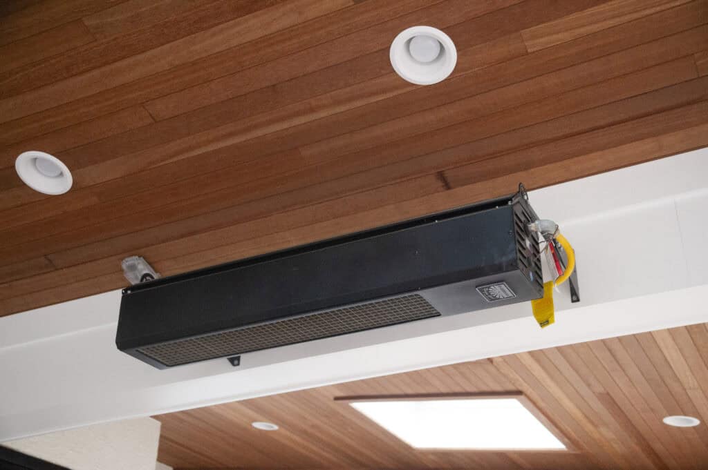 A black ceiling-mounted electric heater with visible wiring is installed on a wooden paneled ceiling with recessed lights and a skylight.