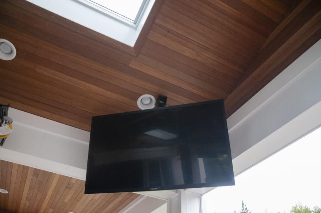 A flat-screen TV is mounted on a wall under a wooden ceiling with recessed lights and a skylight. The TV is turned off, and natural light enters through the skylight.