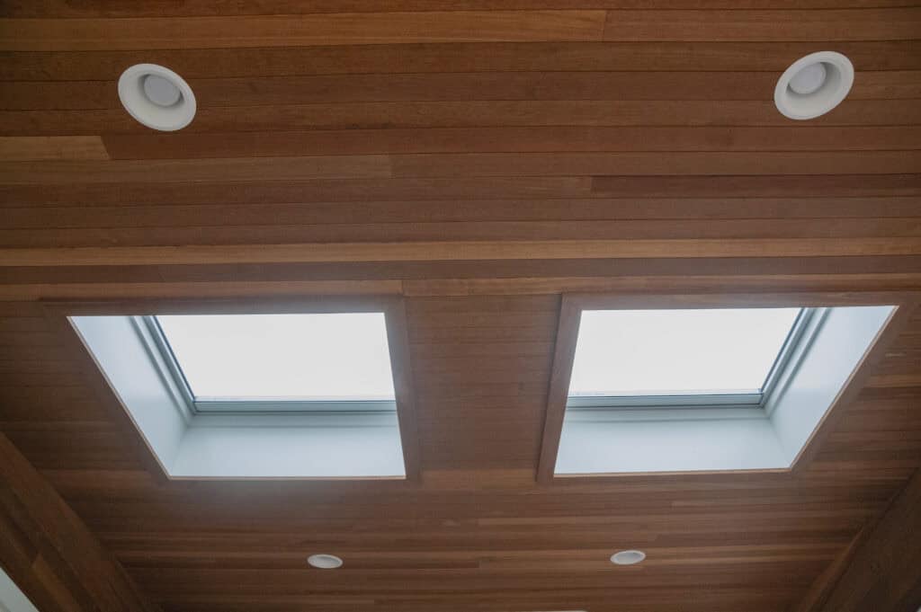 Wood-paneled ceiling with two rectangular skylights letting in natural light, and four round recessed lights evenly spaced around the skylights.